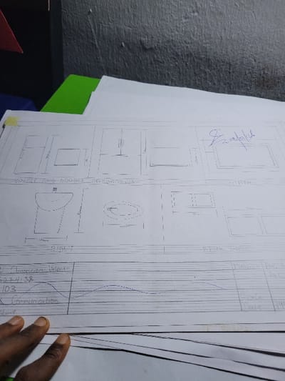 The image displays a close-up, elevated view of a technical drawing sheet laid over a stack of white papers in an indoor setting. The specific location within the city of Puerto López, Colombia, is not visually discernible from the scene. A textured, light gray or off-white wall occupies the upper background.
The central subject is a white sheet of paper containing hand-drawn architectural or technical sketches. The top section of the drawing features "SINGLE AND DOUBLE MEALURIMEMOS" (likely 'MEASUREMENTS') text and depicts various rectangular forms with associated dimensions, possibly representing furniture. To the right, a section labeled "BATH" contains a drawing of a rectangular mirror or frame. Below this, a section labeled "BIACT" includes drawings of a pedestal sink (front view), an oval-shaped fixture (plan view), and two rectangular window-like frames with dimensions. The text "BIRCH WALL" is partially visible in this section.
The bottom portion of the top sheet serves as a title block or data area, partially revealing handwritten text such as "C. Ampencem Valencia," a numerical sequence "5274138," the number "103," and the word "Communication." A wavy line graph extends across this section. Columns labeled "Project," "Date," and "Scale," with a partial date "19XX," are visible on the right. A small piece of yellow adhesive tape secures the upper-left corner of this sheet.
A stack of fanned-out white papers lies beneath the primary drawing. A bright green surface, possibly a folder or desk pad, is visible under the papers on the left. A small, triangular red object is partially visible in the upper left corner, resting on the green surface.
In the lower-left quadrant, a dark-skinned hand is resting on the papers. The fingers are slightly curled, indicating the hand is either holding or simply resting upon the documents. No direct interaction between the hand and the drawings is depicted, but its presence suggests the individual is engaged with the paperwork. The environment appears to be a functional workspace.