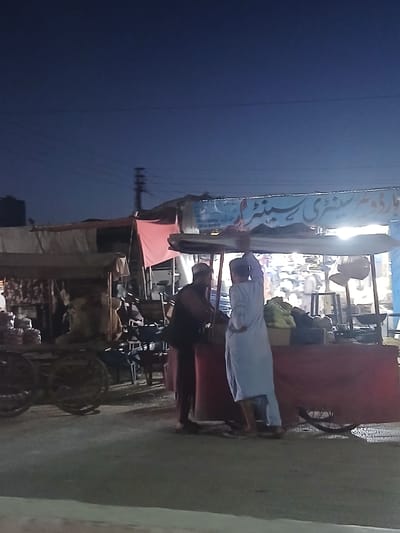 The image shows a street scene at dusk in Kuchlak, Pakistan. The sky is a deep blue, indicating the fading light. A vendor stands in front of a mobile stall selling goods. The stall is covered with a tarp and various items are on display. A customer wearing a white top is interacting with the vendor. A bicycle is parked nearby. The overall impression is of a busy street market.