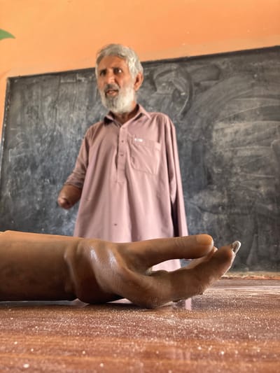 In a classroom setting in Qilla Saifullah, Pakistan, an elderly man with grey hair and a white beard wearing a pink long-sleeved shirt stands in front of a blackboard. His hand is touching a light-brown wooden surface. A layer of white powder covers the surface, including his fingertip. The classroom has a faded orange wall on one side.