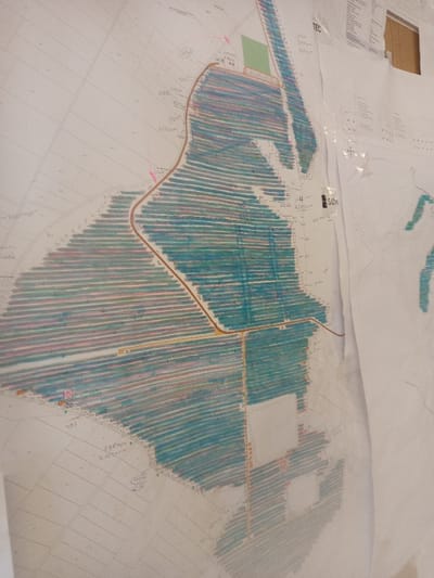 A large, light-colored paper map or cadastral plan is affixed vertically to a wall in Drugan, Bulgaria, secured by transparent adhesive tape visible in the upper right quadrant. The map details a landscape divided into numerous plots, many of which are filled with horizontal marker strokes in teal, blue, and sporadic pink hues. Brown-orange lines, indicative of roads or pathways, intersect and traverse these colored zones. Various handwritten alphanumeric notations and Cyrillic text, including "СУМ" and "ТЕЦ," are present on the map, suggesting measurements, plot identifiers, or project-related information. Several small green rectangles are drawn within certain areas. A separate, uncolored white paper sheet is partially overlaid on the right side of the main map, also secured by tape, and displays a few smaller, irregularly shaped green markings. The surface behind the maps appears to be a light-colored wall. No individuals are visible.