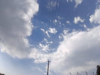 The image depicts a wide-angle view of a cloudy sky, likely taken outdoors. Dominating the upper portion of the frame are numerous white and gray clouds against a vibrant blue sky. Stretching across the middle of the image are multiple dark, thin electrical wires, suggesting the presence of a power line. A single wooden utility pole with insulators stands in the lower central part of the image, supporting the wires. In the lower right corner, the top of a dark-colored tree or foliage is visible, and in the lower left, some indistinct greenery can be seen. The overall scene is a daytime outdoor environment with a focus on the sky and infrastructure. The specific location in Suti, Uzbekistan, is not visually evident from the image itself, but it is the assumed context.