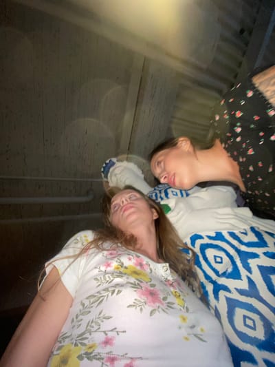 The image shows two women indoors. One woman, in the foreground and on the left, is lying down and looking upwards. She has long, light brown hair and is wearing a white t-shirt with a floral pattern of pink and yellow flowers and green leaves. The second woman is lying down to the right of the first woman and is looking towards the first woman. She has dark brown hair pulled back and is wearing a black t-shirt with a floral pattern of red flowers. Both women are on a surface covered with bedding that has a blue and white pattern. The background consists of a wooden ceiling or wall. The lighting suggests it is nighttime or a dimly lit indoor space. There are no specific city or location details discernible from the image itself that would place it in Solnechnoye, Russia.
