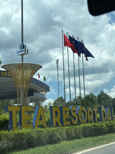 The image shows a large outdoor sign that reads "TEA RESORT DAI" in yellow and blue letters. The sign is set against a backdrop of green foliage and a cloudy sky. To the left of the sign, a tall pole with a circular blue and white logo bearing the word "DOIDEP" is visible. Several flags are flying on poles to the right of the sign, with some appearing to be red and blue. In the distance, a cellular tower can be seen. The scene appears to be a roadside view of an entrance to a resort. This location is Thôn 6, Vietnam.