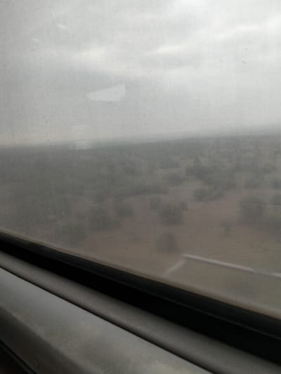 The image shows a landscape viewed through a dirty window. The scene outside is hazy and indistinct, with sparse vegetation scattered across a flat, sandy terrain. Overcast skies contribute to the muted and gray color palette. The window frame is visible in the foreground, adding a sense of enclosure.