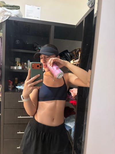 A woman in a navy blue sports bra and black shorts is taking a mirror selfie. She is wearing a dark visor and holding a pink and silver can, possibly an energy drink, to her lips. She is using a green-cased phone. The background appears to be a cluttered closet with shelves filled with bags, clothes, perfume bottles, and miscellaneous items. The closet has dark wooden drawers with silver pulls. A box is seen in the upper left, seemingly sitting on a white surface. The exact location in Villahermosa, Mexico cannot be determined based on the visual information provided.