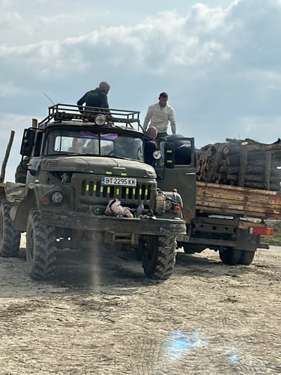 This image, captured in Tsenovo, Bulgaria, depicts two olive-green heavy-duty trucks parked on an unpaved, light-colored dirt surface under an overcast sky. The foreground features a military-style, all-wheel-drive truck, possibly a ZIL-131, equipped with large off-road tires, a metal roof rack bearing auxiliary lights, and a modified front bumper holding a plastic bottle and a light-colored cloth item. The truck displays a Bulgarian license plate "BT 2295 KK." Yellow reflective strips are affixed to the grille and front fenders. Four adult males are present in the scene: one man in a dark jacket is positioned on the roof rack of the lead truck, another man in a light-colored long-sleeved shirt stands on the bed of the rear truck, a third man in a dark shirt is partially visible leaning into the open passenger door of the lead truck, and a fourth person is seated in the driver's seat of the lead truck. The second vehicle, a flatbed truck, is situated behind the first and is heavily loaded with cut logs and wooden planks. Its rear license plate is partially visible, ending in "BOBX". The individuals appear to be actively engaged with the vehicles or their cargo.
