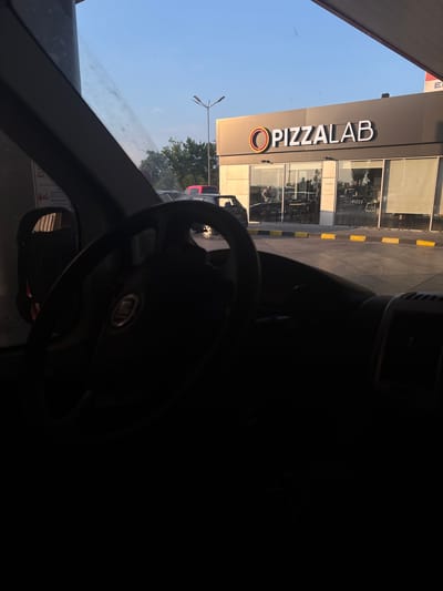 The image provides a driver's-eye view from inside a vehicle, looking towards a commercial establishment in Tsaratsovo, Bulgaria, during what appears to be late afternoon or early evening under a clear blue sky.
The immediate foreground, occupying the lower half of the frame, is dark and shows the interior of the vehicle. A dark, three-spoke steering wheel with a partially obscured central logo (consistent with a Fiat emblem) is prominent on the right. To the left, a dark side mirror and the vehicle's A-pillar are visible. A white sign, possibly a warning sticker inside the vehicle or at a gas pump, shows text including "СТАРТИРАЙ" (Start) and "DO NOT START" in dark letters.
Beyond the vehicle, a paved service area or parking lot stretches towards a single-story commercial building. The paved surface features yellow and black curb blocks separating the driving area from the building's front. Several vehicles are parked in the distance, including a dark hatchback on the far left, a red SUV, and a dark sedan. A streetlight with two luminaires stands tall against the sky on the left, with green trees visible behind it and the parked cars.
The central focus of the background is a modern building with a dark grey upper facade and large, floor-to-ceiling glass windows below. A prominent sign on the facade reads "O PIZZALAB" in white capital letters, with the 'O' stylized as an orange-rimmed circle. Through the glass windows, the interior of the Pizzalab restaurant is visible. Dark tables and chairs are arranged inside, and several individuals are present, appearing to be patrons or staff. One person with dark hair is distinguishable near a counter on the left side of the interior. A small interior sign with "PIZZA" is also visible. To the far right, above the Pizzalab sign, a portion of a red and white canopy or structure with a visible "E" (possibly part of "EL" for a fuel brand) indicates the presence of a gas station adjacent to or co-located with the restaurant.