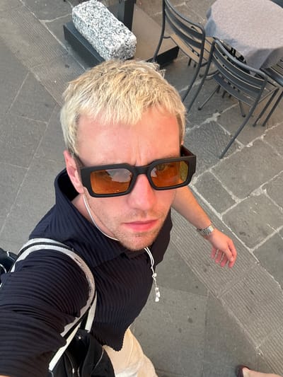 The image captures a high-angle, slightly rotated perspective of a person outdoors in Pisa, Italy, likely a selfie. The main subject is a person with short, bleached blonde hair and light stubble/beard, wearing black-framed sunglasses with reflective orange-tinted lenses. Their face is partially obscured by the sunglasses and the angle. They are wearing a dark blue or black ribbed collared shirt, and a white string, possibly from earbuds, is visible around their neck. On their right wrist, a silver-colored metal watch with a link band is worn. A white strap, possibly from a bag, is visible over their left shoulder.

The setting appears to be an urban street or plaza with grey stone paving. To the left of the person, there's an outdoor cafe area with a small round table covered by a light grey tablecloth and several stacked black metal chairs. A textured, grey speckled stone block is positioned near the person's head in the foreground. In the upper right background, a foot wearing a dark brown or burgundy shoe is visible. The lighting suggests it is daytime, possibly overcast or late afternoon, as indicated by the warm reflections in the sunglasses. There is no visible text in the image.