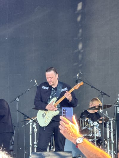 In Bolgar, Russia, a male guitarist on stage, wearing a black jacket with "IRAN" and "JILLEN" patches and dark pants, plays a light green electric guitar. He is positioned center-left. To his right, a drummer with blond hair, wearing a black shirt, plays a drum kit. The background is a dark, slightly textured surface. A person's hand, wearing a watch, is in the foreground, holding a purple smartphone to take a picture of the performers. The lighting suggests an outdoor daytime event.