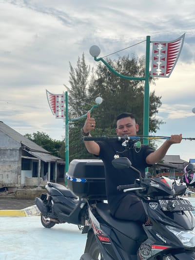 This is a portrait-oriented, slightly tilted photo of a man holding a fishing rod. He is sitting on a black motorcycle, which is parked next to a light-colored wall. The man is wearing a black t-shirt and a watch. He is smiling and giving a thumbs-up gesture. The motorcycle has various stickers and a license plate that reads "BL3 NAC 01-22". In the background, there are trees and a cloudy sky. Two decorative flags with a red and white sawtooth pattern are visible, mounted on green poles. A building with a weathered appearance is also in the background. The overall scene suggests a casual outing, possibly near a coastal area given the general ambiance and the fishing rod. The time of day appears to be daytime, with diffused light from the overcast sky. The location is identified as Lancok, Indonesia.