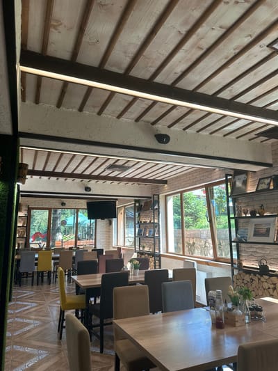 The image depicts the interior of a restaurant in Sarnitsa, Bulgaria. The space features a rustic design with wooden beams and white-washed wooden planks on the ceiling, complemented by exposed brick walls and large windows offering views of greenery. The dining area is furnished with several tables and chairs of various colors, including grey, yellow, and beige. On the tables, condiments, cutlery, and floral arrangements are visible. Decorative shelves are placed against the walls, displaying pictures and other items. A television screen is mounted on one of the walls. The flooring is tiled with an intricate pattern. The overall atmosphere suggests a well-lit and inviting dining establishment.