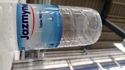 A horizontal, partially-filled, clear plastic bottle is centered in the foreground. The bottle's label is white and blue, prominently displaying "Jazmyne" in blue lettering, with "Table Water" and "Refreshing" also visible, alongside a blue water drop icon. Condensation droplets adhere to the exterior surface of the bottle. The background is out of focus, revealing an indoor setting characterized by structural components: horizontal metallic bars, angled dark metallic beams, and corrugated or textured surfaces consistent with a roof or ceiling. No individuals, actions, or specific identifying landmarks for a precise location within Bwari, Nigeria, are discernible.
