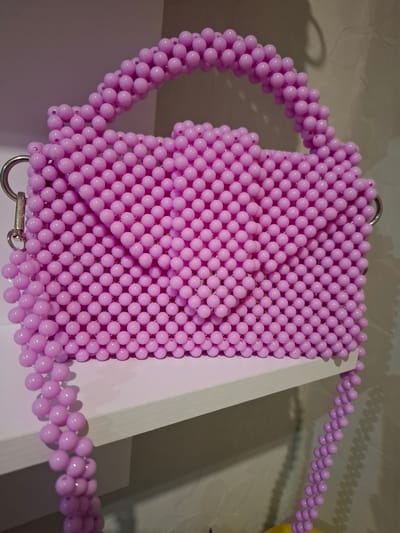 A close-up, eye-level shot shows a light purple beaded handbag with a silver clasp and strap hardware. The handbag is placed on a white shelf against a wall with a subtle textured pattern. The lighting is diffused, casting soft shadows. The background appears to be an indoor setting. No people or other objects are clearly visible.