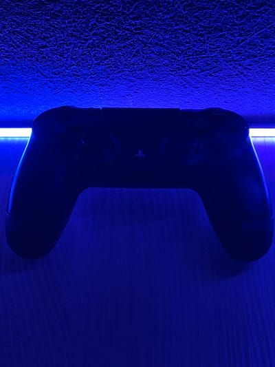 The image shows a black video game controller resting on a wooden surface, illuminated by blue light. Above the controller, a textured ceiling is visible, also bathed in blue light. The controller is centered in the frame, with its handles extending to the left and right. The PlayStation logo is faintly visible on the face of the controller. A bright white light source runs horizontally across the top of the controller, casting a strong glow. The wooden surface appears to have a visible grain pattern. The overall impression is that of a gaming setup in a room, possibly a bedroom or entertainment area. No people are present, and no specific location in the city of Ардонь, Russia, can be identified from the image.