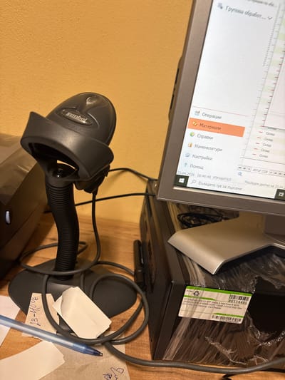The image depicts a cluttered indoor workspace in Glava, Bulgaria, featuring office equipment. No people are visible.
The central objects include a computer monitor displaying a software application interface in Bulgarian. Text on the screen identifies sections like "Операции" (Operations), "Материали" (Materials), "Справки" (Reports), "Номенклатури" (Nomenclature), "Настройки" (Settings), and "Помощ" (Help). A counter indicates "207 of 1328" records, and the last access date is partially visible as "Последен достъп на 23.04.2". The system tray shows the date and time as "23.04.2026 16:46:46" (April 23, 2026, 4:46 PM) and identifies the user as "УПРАВИТЕЛ" (MANAGER), alongside a search bar with the prompt "Въведете тук за търсене" (Type here to search).
Next to the monitor, a desktop computer, an "HP EliteDesk 800 G1 SFF Grade A," is visible, still partially encased in protective plastic. A label on its side lists specifications: "Intel Core i5 4570, 3200MHz 6MB, 8192MB DDR3, 128GB 2.5 Inch SSD Slim, DVD-RW Sim Desktop Card Reader." Other details on the label include "80114495," "ISO 9001," and a serial number "CZC4040VBR."
Below the monitor, a black "Symbol" brand barcode scanner is positioned on its stand. Various black cables are scattered across the wooden desk surface. A blue pen and several pieces of paper, one with "23-40UE" handwritten on it, lie near the scanner's base.
The setup, including the computer, software in Bulgarian, and barcode scanner, suggests an active workstation used for tasks such as inventory management, data entry, or administrative processing within an office or retail environment. The time displayed indicates the capture or last activity date.