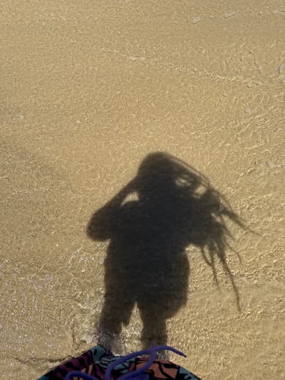 The image shows a shadow of a person, possibly taking a photograph, standing in shallow water on a beach. The sand is light beige and visible through the clear water. Part of a purple and blue bathing suit is visible on the right side of the image. The location is likely a beach in Pájara, Spain, as specified in the prompt.