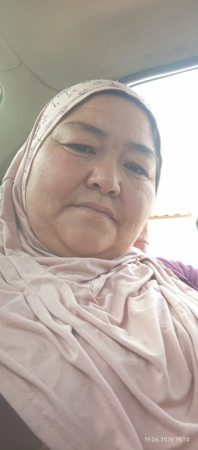 This is a close-up, medium shot selfie of a woman wearing a pink hijab and a matching pink garment. The woman's face is the primary subject, with her eyes looking towards the camera. Her skin has visible texture, and she has a neutral expression. The hijab is adorned with small, scattered beads in various colors, including pink and white. The lighting suggests it's daytime, possibly outdoors or near a window, with bright light coming from the right side of the frame. The background is blurred and out of focus, appearing to be an interior space, possibly a car given the ceiling material visible in the upper portion of the image. A small portion of a purple garment is visible on the right side of her shoulder. In the bottom right corner, there is a timestamp: "19.06.2026 15:10". The woman appears to be in Чоң-Гара, Kyrgyzstan.