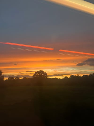 The image captures a sunset over a silhouetted treeline in Forchheim, Germany. The sky is a dramatic gradient of colors, transitioning from a deep blue at the top to vibrant oranges and yellows closer to the horizon. Two distinct contrails, one bright orange and the other white, streak across the sky from left to right, suggesting aircraft activity. Below the sky, a dark silhouette of trees forms the horizon, with a field or open space visible in the foreground, also rendered in dark, indistinct tones due to the low light. The overall scene is one of natural beauty enhanced by atmospheric phenomena and the remnants of human activity.