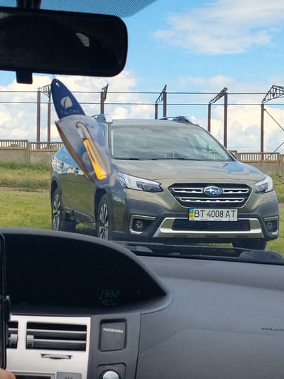 The image shows a dark-colored Subaru Outback parked outdoors, viewed from inside another vehicle. A car air freshener, partially obscuring the Subaru's windshield and front grill, hangs from the rearview mirror of the vehicle taking the photo. The Subaru's license plate reads "BT 4008 AT" and appears to be a Ukrainian plate. The car is parked on a grassy area with a concrete barrier and industrial-looking structures in the background. The sky is blue with white clouds. Inside the vehicle where the photo is taken, a digital display shows the time as "13:32" and a speed of "45.7 km/h". The dashboard also features air vents and a button labeled "PASSENGER". The presence of a Ukrainian license plate on the Subaru suggests it might be a vehicle from Ukraine, but the specific location in Colonița, Moldova is not directly indicated by any visual cues in the image itself.