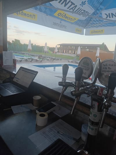 The image depicts a view from a bar counter looking out towards a swimming pool area. In the foreground, there are three beer taps, a laptop displaying a spreadsheet, a paper cup, a roll of tape, a smartphone, and some documents. The laptop is open and positioned on the bar. Behind the bar, umbrellas and lounge chairs are visible around a swimming pool. In the background, a two-story building with balconies and several windows is situated. The sky is overcast with some visible clouds. There are no people clearly visible in the image. The scene suggests a commercial establishment, likely a bar or restaurant with a pool, in Shkurupiivka, Ukraine. The overall impression is one of a quiet, possibly late afternoon or early evening setting.