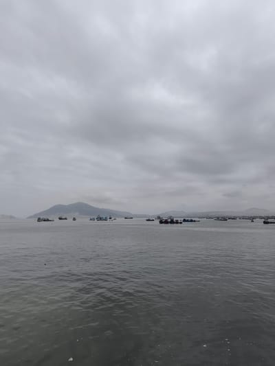The image depicts a seascape under a heavy, overcast sky, likely taken in or near Chimbote, Peru. The sea is gray and choppy, reflecting the dull light. Numerous boats, likely fishing vessels, are scattered across the water. In the distance, a mountainous landmass is visible. There is no evidence of human activity in the immediate foreground. The overall scene conveys a sense of gloom and maritime activity.