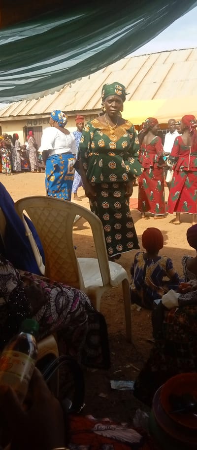 An outdoor gathering is depicted in Kasuwan Magani, Nigeria, beneath a partially visible dark green fabric canopy providing shade. The ground consists of exposed reddish-brown earth. In the mid-foreground, a woman stands centrally, facing forward, dressed in a dark green patterned traditional outfit featuring gold crown motifs, a matching headwrap, a prominent gold necklace, and earrings. To her left, a woman in a white blouse and blue patterned wrapper faces away from the camera. To her right, several women are visible wearing matching red patterned outfits and headwraps. A young child, dressed in a blue and yellow patterned garment and a red head covering, is seated on the ground in the mid-foreground, facing away. In the immediate foreground, a hand holds a small plastic bottle labeled "Light Lemon Lime." Partial views of seated individuals and a white plastic chair are present. On the far right foreground, a stack of orange and purple plastic bowls with a spoon rests on the ground. The background features a light-colored building with a corrugated metal roof, displaying painted text including "MODERN" and "BEAUTY." A bright, hazy sky is visible above the building.