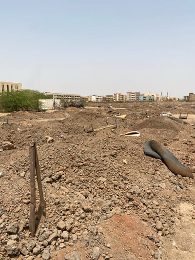 The image shows a large, dirt lot in Khartoum, Sudan, located at 15.528° N, 32.571° E. The lot is filled with rubble and rocks, with a few scattered pieces of debris. There are some buildings in the background, suggesting that the area is likely undergoing urban development. The scene is barren and desolate, with no people or animals present. The sun is shining brightly and the sky is clear. There is a wooden stick sticking out of the ground in the foreground.  A discarded black tarp lies on the ground.  The image is taken from a perspective that looks out over the lot toward the buildings in the background.  The scene is a typical example of urban sprawl and the rapid development that is taking place in many developing countries.