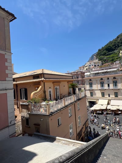 This is a photograph taken on a sunny day in Pogerola, Italy. The scene is a bustling street with many people walking around. Buildings line the street, some of which appear to be shops or restaurants with outdoor seating. The architecture suggests a Mediterranean setting, with stucco buildings and tiled roofs. A steep, tree-covered hillside rises behind the buildings, adding to the dramatic landscape. The time of day appears to be midday, given the bright sunlight and the shadows cast by the buildings and umbrellas. The atmosphere seems lively and energetic, with many people enjoying the pleasant weather and the vibrant street life. There is no visible text in the image.