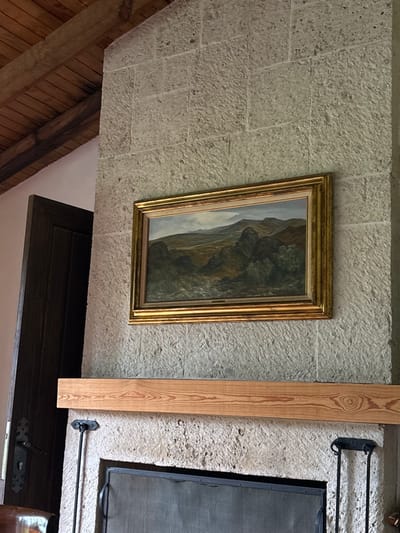 This image captures an interior architectural view in Huasca de Ocampo, Mexico, centered on a stone fireplace. The dominant structure is a fireplace constructed from light-grey, textured, rectangular stone blocks laid in horizontal courses. A light-colored wooden mantelpiece, featuring distinct wood grain, spans horizontally above the fireplace opening. Above the mantel, a rectangular landscape painting is mounted on the stone wall. This artwork is framed in an ornate gold frame and depicts a rugged, mountainous terrain with varied green and brown foliage under a muted sky. Below the mantel, the fireplace opening contains a dark metal mesh screen. To the right of the fireplace opening, two black metal fireplace tools are suspended. To the left of the fireplace, a dark-stained wooden door or archway with vertical paneling and visible dark metal hardware is partially visible. The upper left corner of the image shows a wooden ceiling composed of dark, exposed beams and lighter wooden planks. No individuals are present in the scene.