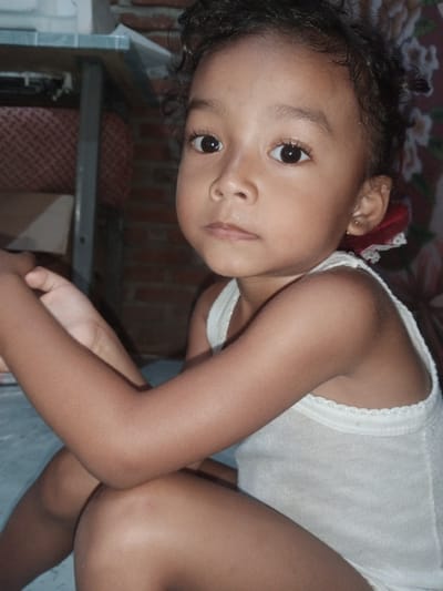 A young girl with dark, curly hair is seated indoors, looking towards the camera. She is wearing a white tank top. Her expression is neutral. She is positioned on a light blue surface, possibly a floor. To her left, a table with a grey metal frame and a patterned surface is visible, behind which is a wall made of reddish-brown brick. To her right, a floral patterned fabric is visible. The lighting suggests an indoor setting, possibly during the daytime. No visible text is present in the image. The location context is Cotseutui, Indonesia.
