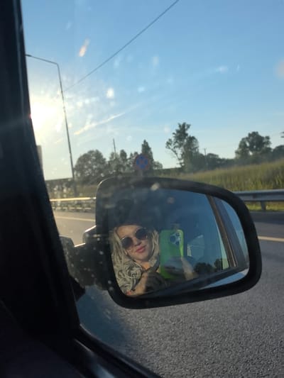 A woman with blonde curly hair and sunglasses takes a selfie in a car's side mirror. She is holding a bright green smartphone in her right hand. The reflection shows her face from the chest up, with her hair framing her face and her lips slightly parted. The background in the mirror is blurred but suggests a roadside environment with a metal guardrail, trees, and a bright sky. The car appears to be on a road with asphalt. The image appears to be taken during daylight, likely late afternoon or early morning, given the angle of the sun. There is no information available to determine the specific location in the city of Krasnyi Oktyabr', Russia.