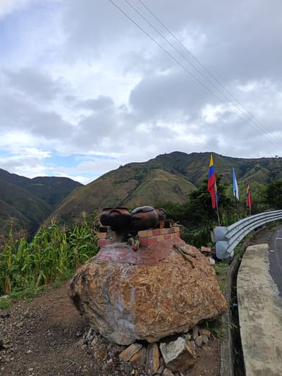 The image depicts a roadside scene in El Cisne, Ecuador. The foreground features a large, textured boulder supporting a brick structure with two dark-colored pots on top. Corn stalks grow on the left side of the boulder. Three flags—two Colombian and one light blue—stand behind the pots, next to a metal guardrail and a concrete road edge. In the background, rolling green hills stretch towards the horizon under a cloudy sky with a patch of blue. Power lines traverse the upper portion of the frame.