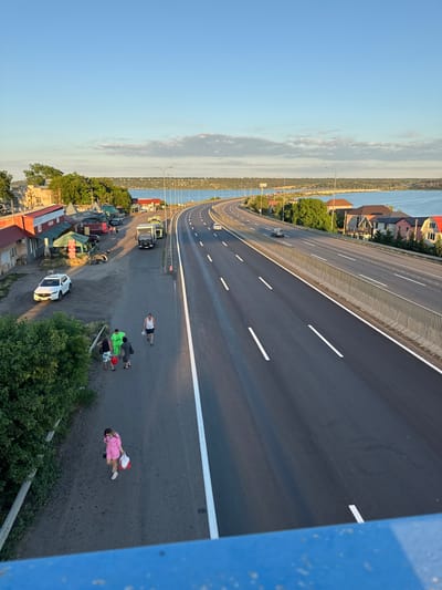 The image depicts a view of a highway running alongside a body of water, likely a river or reservoir, with a town or village on the left bank. The scene is set in Yelyzavetivka, Ukraine.

On the left side of the highway, there is a row of small buildings, some with red roofs, resembling shops or market stalls. A white SUV is parked in front of one of these buildings. Further down the road, a group of people are walking. One person is wearing a bright green outfit, and two others appear to be a couple. Another individual, a woman in a pink outfit, is walking further down the sidewalk carrying shopping bags. Several parked trucks and cars are visible near the buildings.

The highway itself is a multi-lane road with clear white markings. Cars are driving on the highway in both directions. A concrete barrier with a metal railing separates the highway from the sidewalk area.

In the background, across the water, there are hills and trees. The sky is a clear blue with a few scattered clouds, suggesting it is daytime, possibly late afternoon or early evening due to the soft lighting. The overall impression is a semi-urban or rural setting where a major road passes through a populated area near a natural water feature.