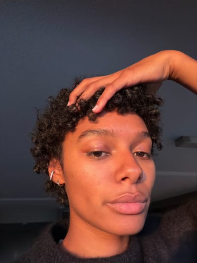 The image presents a close-up portrait of an individual with dark, curly hair, oriented on their side. The person's face is illuminated by a strong, warm light source from the left, highlighting their forehead, cheekbones, nose, and lips, and creating a soft glow on the skin. Their dark eyes are open, looking upwards, and they have full, light pink lips.

One arm is raised, with fingers gently resting on the person's head near their dark, tightly coiled hair. A small, sparkling hoop earring is visible in the left earlobe. On the right side of the frame, a dark, fuzzy or knitted garment is partially visible, suggesting a sweater or similar clothing. The background consists of a plain, dark blue-grey wall, creating a stark contrast with the illuminated face. In the top right corner, a faint, lighter-colored rectangular object, possibly an architectural feature or light fixture, is barely discernible.

The overall mood is calm and introspective. There is no discernible activity taking place other than posing for the photograph. The warm lighting suggests either natural light during golden hour or specialized indoor lighting. No specific outdoor or location cues are visible that would link directly to Champs-sur-Marne, France, beyond the indoor setting. There is no visible text in the image.