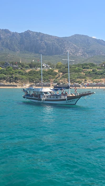 A tour boat named "SHAKKI BOAT TOUR" is anchored in the turquoise waters off the coast of Karavas, Northern Cyprus. The boat is white with brown trim and has two masts. Several people are visible on the deck of the boat, some sitting and others standing. In the background, a sandy beach is lined with trees and mountains. A parking lot is visible behind the beach with cars parked. The sky is clear and blue.