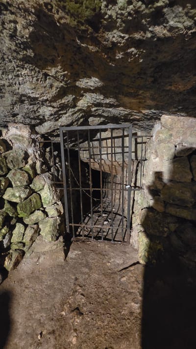 The image depicts a metal grate secured over a dark, narrow opening within a stone structure, likely in Altura, Spain. The opening is surrounded by rough, uneven stone walls and rubble. The grate is constructed of rectangular metal bars arranged in a grid pattern. The overall environment suggests a cave or underground chamber. The lighting is dim, obscuring details further into the opening. No people are visible.