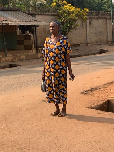 This image depicts a mature adult female standing on a dirt roadside in Otuocha, Nigeria. She is positioned slightly left of center, gazing towards the left. The woman, appearing to be of African descent, wears a dark blue, V-neck, ankle-length dress patterned with yellow and white leaf-like motifs, and a dark blue headwrap covering her hair. Her skin is dark, and her facial expression is neutral. Her left hand holds a dark, possibly plastic, bag, while her right hand grips a small, light-colored rectangular object, possibly paper or a wrapper. She wears open-toed sandals on her feet.
The immediate foreground consists of reddish-brown compacted dirt with an irregular, shallow drainage ditch visible to the right of the woman. Behind her is a paved road. In the mid-ground to the left, a small, single-story structure with a corrugated metal roof and walls featuring a brown and beige checkerboard tile pattern is visible. To the right, a light-colored concrete wall, approximately head-height, topped with coiled barbed wire, runs horizontally across the background. Integrated architectural pillars are visible along this wall. Behind the wall, a tree with bright green and yellow foliage is present. The scene is illuminated by direct sunlight, casting shadows to the right of the subject, indicating daytime conditions.