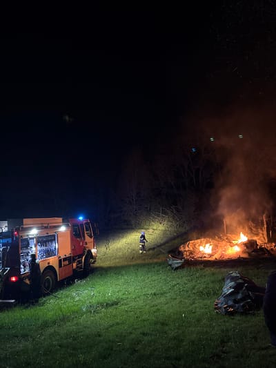 The image captures a nighttime scene in Dolno Drianovo, Bulgaria, dominated by a burning fire and the presence of emergency services.
In the foreground, a large fire is ablaze, emitting bright orange and yellow flames and a significant amount of smoke, which glows orange from the firelight. The fire appears to be consuming a pile of debris or other material, leaving charred remnants. Near the fire, a firefighter stands, illuminated by the flames and other light sources. This individual is wearing a protective suit, blue on the top half and yellow on the bottom.
To the right of the scene, a red and white fire truck is parked on grassy ground. Its emergency blue lights are active, casting a blue glow on the immediate surroundings. The truck's compartments are open, revealing various pieces of equipment and hoses. A hose can be seen extending from the truck towards the area of the fire, indicating active fire suppression or monitoring. Another person, likely a firefighter, is visible near the back of the truck. The side of the fire truck features partial text, including "ПОЖАРНА" (Fire Department) and possibly "ДЕЖУРЕН".
The entire scene is set outdoors at night. The ground is covered in grass, and bare tree branches are visible against the dark sky in the background, partially obscured by smoke. The only significant illumination comes from the intense flames of the fire and the spotlights emanating from the fire truck, casting harsh shadows and highlighting areas of interest. The overall impression is one of an ongoing emergency response to a fire incident.