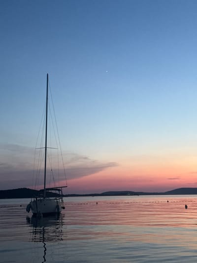 A sailboat is anchored in calm water at dusk. The sky is a gradient of blue to pink, with a few wispy clouds. The water reflects the colors of the sky. A string of buoys is visible in the distance. The sailboat has a tall mast and rigging. It appears to be a calm evening in Grad Šibenik, Croatia.