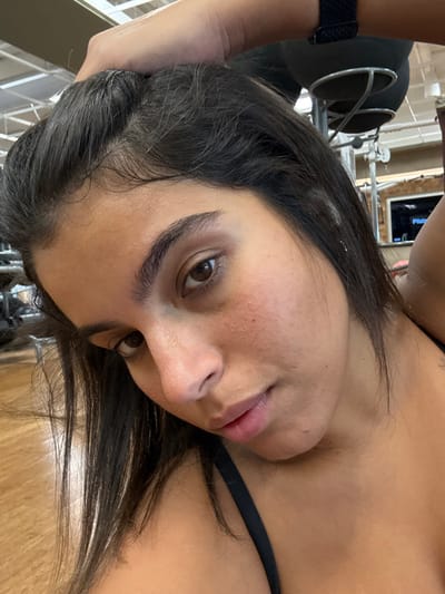 A young adult female is depicted in a close-up, chest-up view within an indoor gym or fitness center. She has dark brown hair and brown eyes, with visible perspiration on her forehead and cheeks. She is wearing a black sports bra, with one strap visible on her left shoulder. Her right arm is raised, with her hand resting on the back of her head, appearing to push her hair back. She maintains direct eye contact with the camera. The background is blurred, revealing gym equipment including metallic frames and a black padded seat on the upper right, consistent with a Pilates reformer or similar apparatus. A rectangular screen in the background displays white text, partially legible as "Pilates." The floor in the lower left foreground is light-colored wood. The specific location within Plainsboro Township, United States, cannot be determined from the visual evidence.