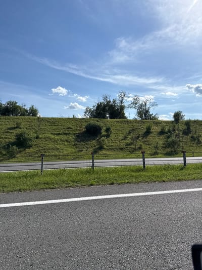 This media file depicts a rural roadside scene in Belarus under a bright, partly cloudy sky. The composition is dominated by a sloping grassy embankment on the left, dotted with trees and bushes, and a paved road on the right with a white line marking its edge. A thin, paved path runs parallel to the road, separated by a low fence with vertical posts.
The time of day appears to be daytime, likely late morning or early afternoon, given the bright sunlight casting shadows. The weather is pleasant, with scattered white clouds against a clear blue sky, suggesting a warm, sunny day. The location is hinted at by the presence of what appears to be a highway or main road with a bike path, bordered by natural, undeveloped land. The overall impression is one of tranquility and an open, natural environment. No people or specific activities are clearly visible. The image is oriented vertically, with the sky occupying the upper left portion and the road and embankment filling the rest.