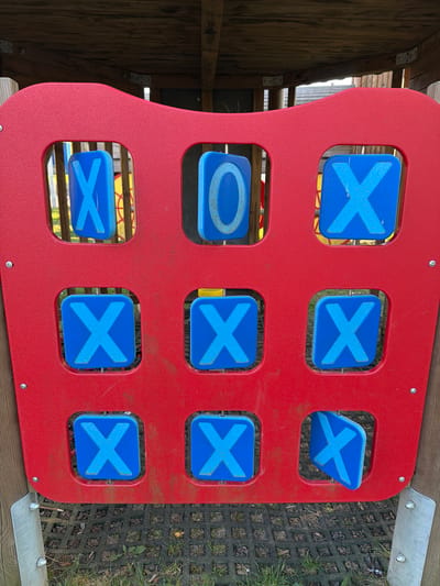 This image displays a red outdoor Tic-Tac-Toe (Noughts and Crosses) game panel, likely situated in a playground in Niederanven, Luxembourg. The panel is made of textured, red plastic, showing some signs of wear and dirt. It features nine square cutouts, each containing a rotatable blue plastic piece. Most of these pieces are currently displaying a light blue 'X' symbol. Specifically, eight 'X' symbols are visible across the board, arranged in various rows and columns. One blue piece in the middle-left position shows a light blue 'O' symbol. A piece in the top-right corner is partially turned, showing its edge, but still displays an 'X'.
The arrangement of 'X' symbols indicates multiple winning lines for the 'X' player, including horizontal wins in the top and bottom rows, vertical wins in the second and third columns, and diagonal wins. The game board is attached to a wooden structure on the left, possibly part of a playhouse or climbing frame. To the right, a dark grey grid-patterned ground surface, possibly rubber matting or concrete pavers, is visible with grass growing through. Hints of other yellow and blue playground equipment can be seen in the background through the panel's openings. The scene is illuminated by natural daylight, without strong shadows, suggesting an overcast day. No text is visible, only the game symbols.