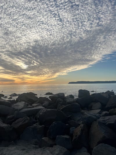 A sideways view of a body of water meeting a textured sky. A pile of dark gray rocks fills the right side of the frame. The water reflects the sunset colors, transitioning from orange to light blue. Above the water, the sky is filled with cumulus clouds, creating a rippled pattern with sunlit edges. The clouds shift to a textured, almost grainy cloud formation towards the left side.