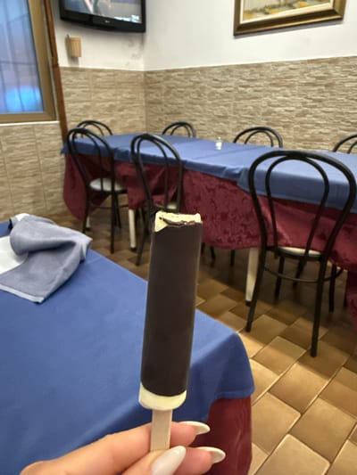 In a dimly lit indoor space, possibly a restaurant or dining area in Sesto San Giovanni, Italy, a person's hand with manicured fingernails holds a chocolate-covered ice cream bar. The ice cream bar has a dark chocolate coating over a lighter interior and is attached to a wooden stick. The background reveals several round tables covered with blue cloths and red tablecloths underneath. Black metal chairs with curved backs are arranged around the tables. The walls are tiled with a textured, light brown material. A television is mounted on the wall in the upper left corner. The floor is tiled with square, light brown tiles.