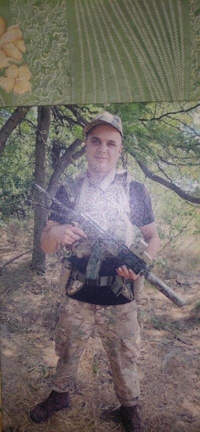 A full-body photograph depicts a man dressed in camouflage attire, including a tactical vest, pants, and a baseball cap. He stands in a wooded area, holding a rifle with both hands. The man appears to be in his late 20s or early 30s, with short dark hair and a neutral facial expression. The background consists of trees and dry vegetation, suggesting an outdoor, possibly rural, setting. The image has a slightly grainy texture, which may be due to the photographic medium or resolution. The location is identified as Ketrysanivka, Ukraine.
