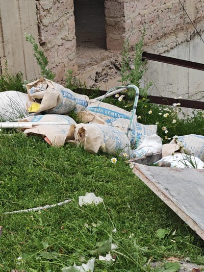 The image shows a pile of cement bags on a grassy area next to a partially constructed or dilapidated stone and concrete structure. The cement bags are mostly light brown paper with blue lettering. Some bags have visible text including "URMIA CEMENT", "M-500", and "EXPORT". There is also a white plastic pipe and some scattered debris, including a piece of metal sheeting in the foreground. Small white daisies are interspersed among the bags and grass, suggesting a sunny outdoor setting. Green weeds and grass are growing around the materials, indicating the location is likely outdoors and possibly abandoned or under construction. The stone structure appears to be made of rough-hewn blocks, with a rough concrete section to the left and a more refined, possibly eroded, stone section to the right. A dark metal bar runs horizontally across the image in the middle ground, attached to the wall. The overall scene suggests a construction site or a storage area for building materials in an outdoor, possibly rural, setting. The time of day appears to be daytime, given the lighting. No people are visible in the image.