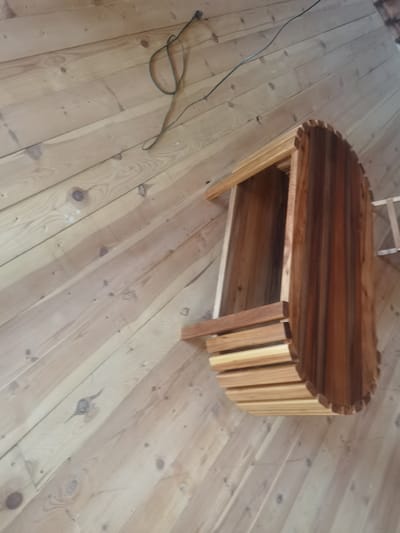 A wooden shelf with a curved, slatted front is mounted on a sloped wooden wall. A black electrical cord is draped across the wall near the top of the frame. The wall is constructed of light-colored wooden planks, running diagonally from the bottom left to the top right. The shelf itself is made of darker wood, with a curved front composed of horizontal wooden slats. The overall scene appears to be an interior of a room with a wooden ceiling and walls.