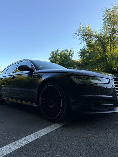 A dark-colored sedan is parked on an asphalt surface, indicated by a white parking line. The car is positioned at an angle, with its front end and passenger side prominently visible. The front of the car features a sleek headlight design and a large grille. The wheels are dark, multi-spoke rims. Trees with lush green foliage are visible in the background, casting shadows on the car and the ground. The sky is clear and blue, suggesting a sunny day. The lighting and shadows indicate that the sun is high in the sky, likely mid-day. The overall scene depicts a car parked outdoors in what appears to be a residential or parking area, given the presence of a parking line and the backdrop of trees. No people are visible in the image. The image is oriented vertically, with the top of the car's front end appearing on the left side of the frame.