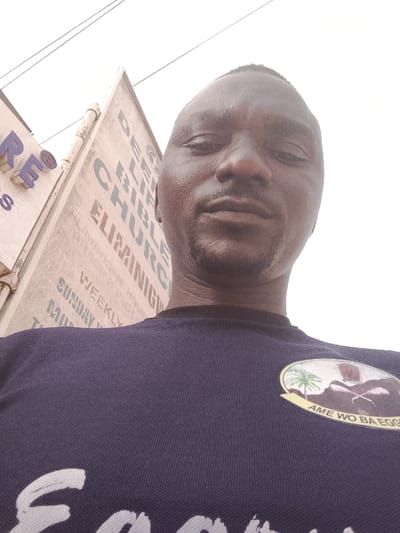 A man takes a selfie outdoors. He wears a navy blue t-shirt with an emblem featuring a palm tree, a figure, and the words "AME WO BA EGG". Behind him is a building with signage reading "BIBLE CHURCH".  Other text on the sign includes "ELIMINIG," "WEEKLY," "SUNDAY", and "MON". Power lines are visible in the upper portion of the image.