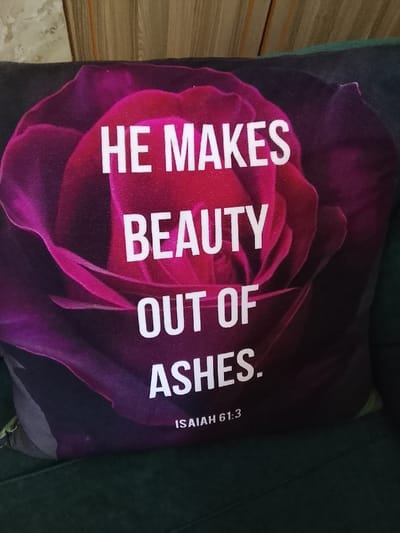The scene is an interior setting, located in Gatongora ward, Kenya. A rectangular throw pillow is centrally positioned, featuring a large magenta and dark pink rose graphic against a dark background. White, sans-serif capital letters on the pillow read "HE MAKES BEAUTY OUT OF ASHES." Below this text, also in white capital letters, is "ISAIAH 61:3." The pillow rests on a dark green upholstered surface, consistent with a sofa or chair. Behind the pillow, a light brown, vertically textured surface is visible, likely a wall or wooden panel. No individuals, animals, or active events are present. The lighting is ambient and indicative of an indoor environment.